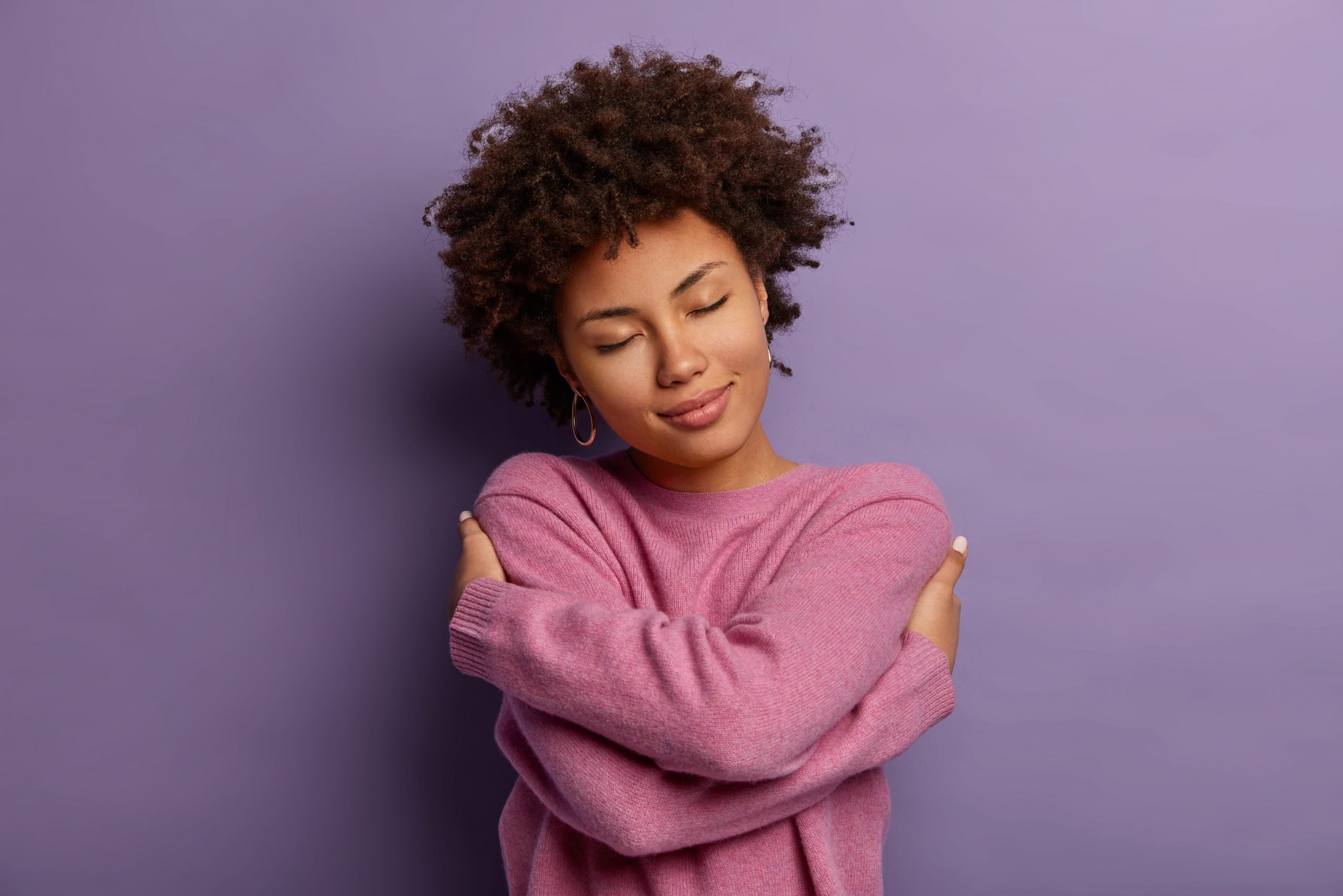 Romantic woman embraces herself, keeps arms crossed over body, touches shoulders, enjoys coziness, wears sweater, tilts head, has high self esteem, loves herself, isolated on purple background